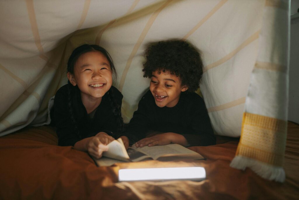 Two children smiling and reading a book in a cozy blanket fort indoors.